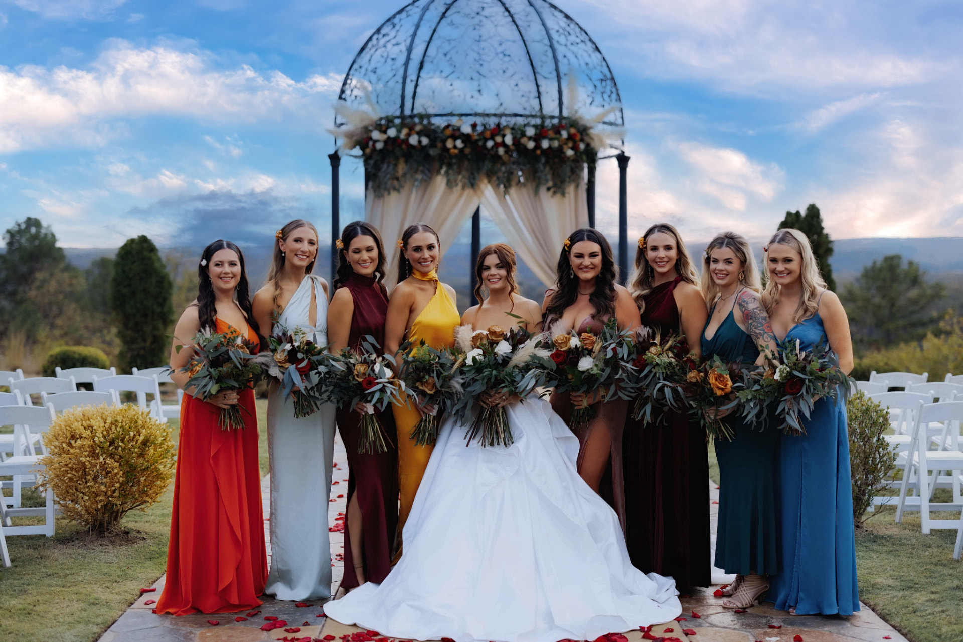 Bride with her bridesmaids in colorful gowns beneath a floral gazebo at sunset